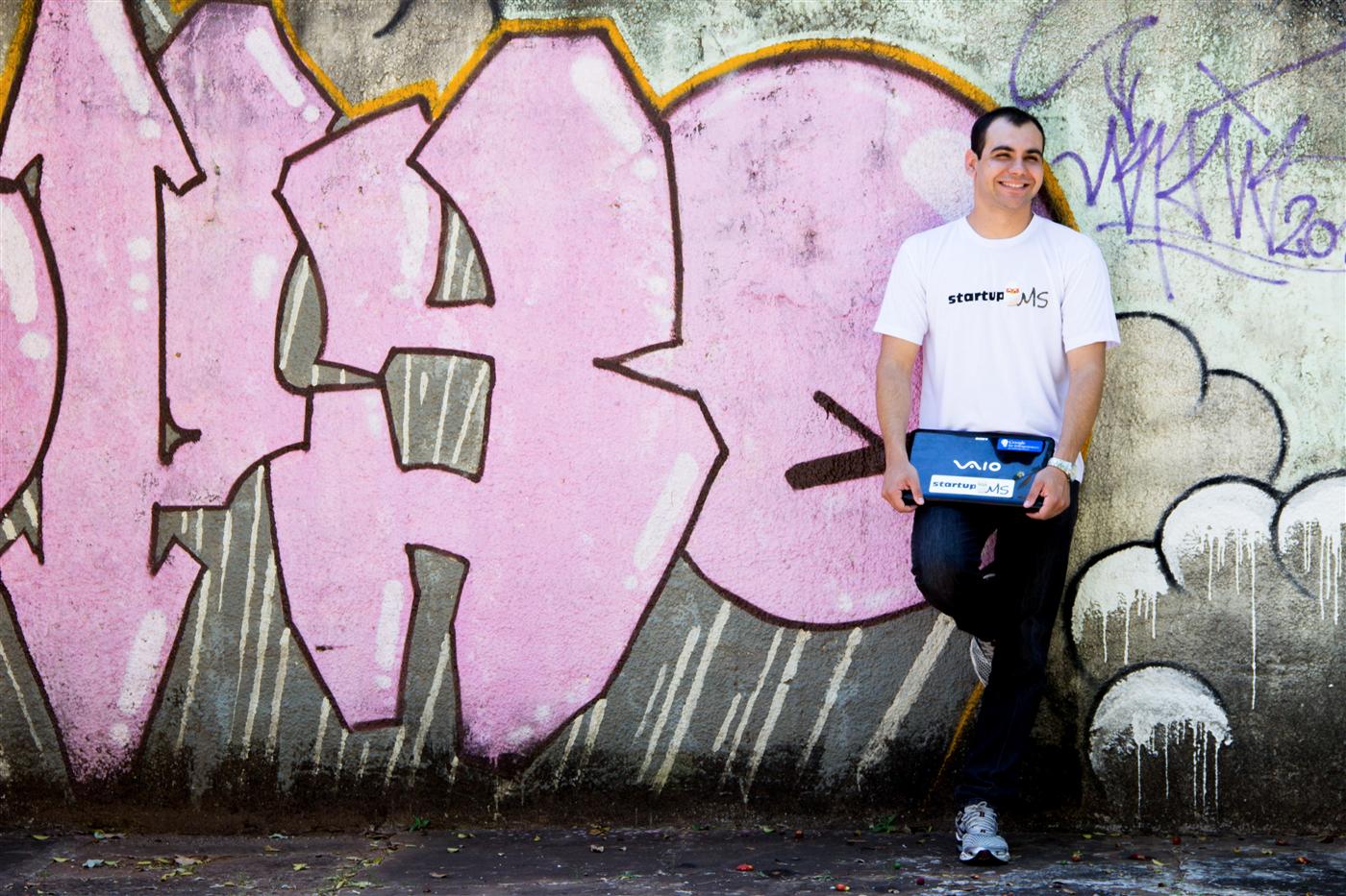 Rodney sorrindo com camiseta e notebook da StartupMS diante de um muro grafitado